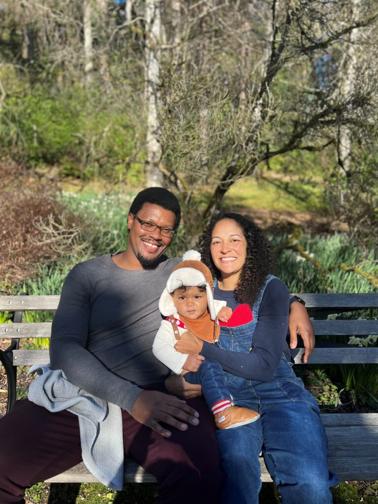 A beautiful Black family consisting of two parents (named Dion and Evelyn) sitting on a bench in front of greenery, holding their 11-month-old son (Danin).